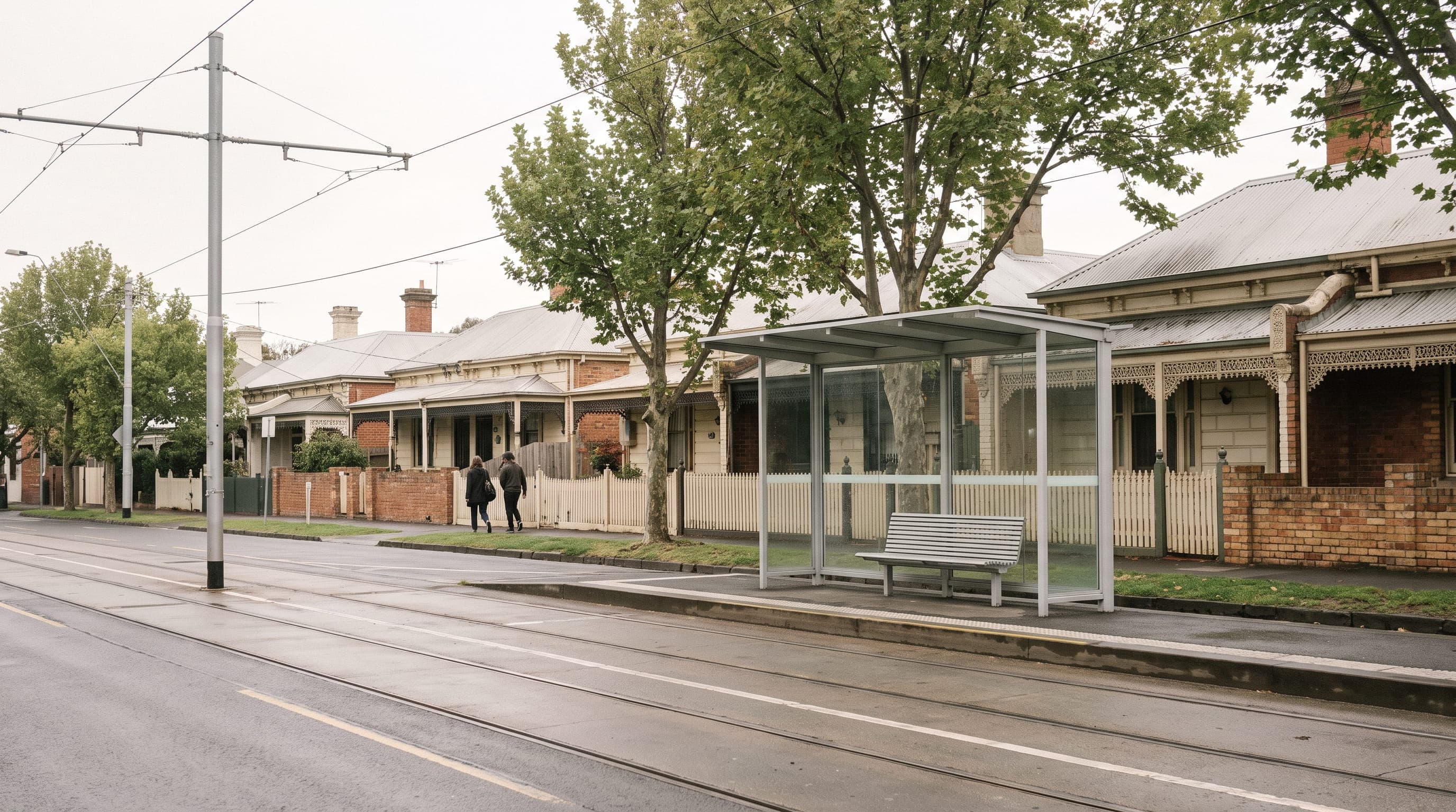 Two people walking along a suburban street past a tram stop, with Victorian cottages and plane trees behind them.